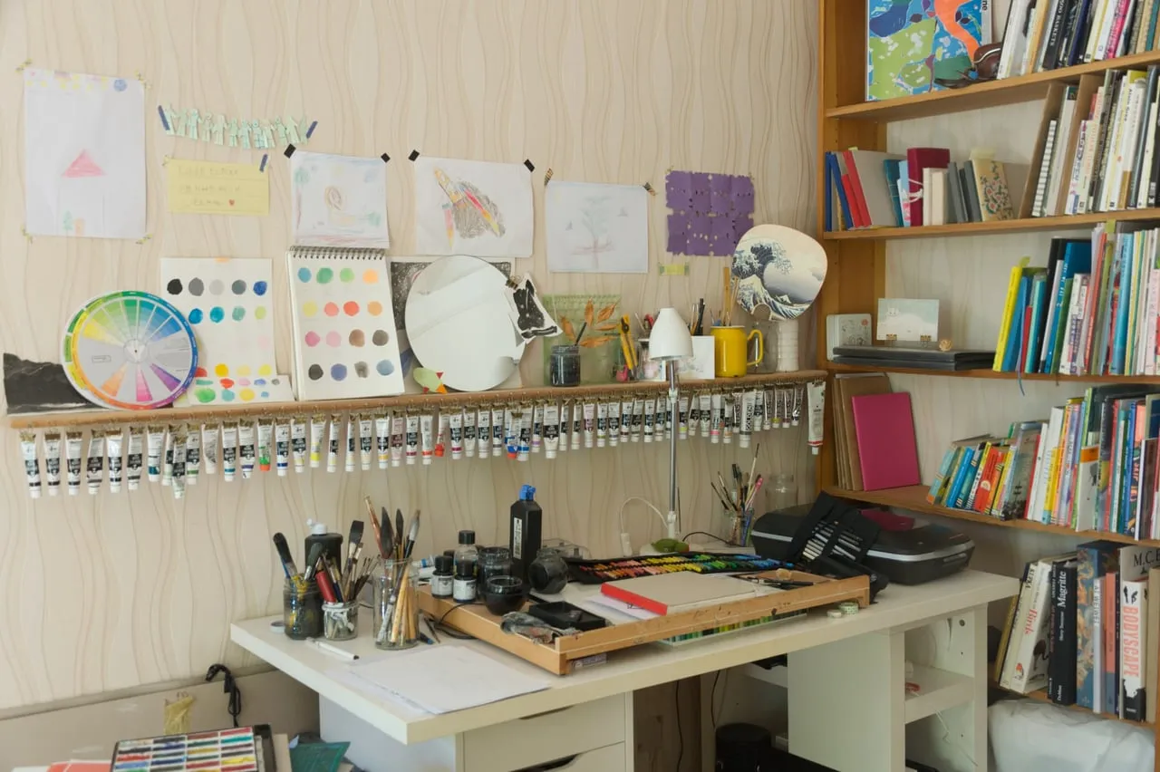 Risa’s studio desk beneath shelves of books, with paint tubes, brushes and sketches arranged along the wall.