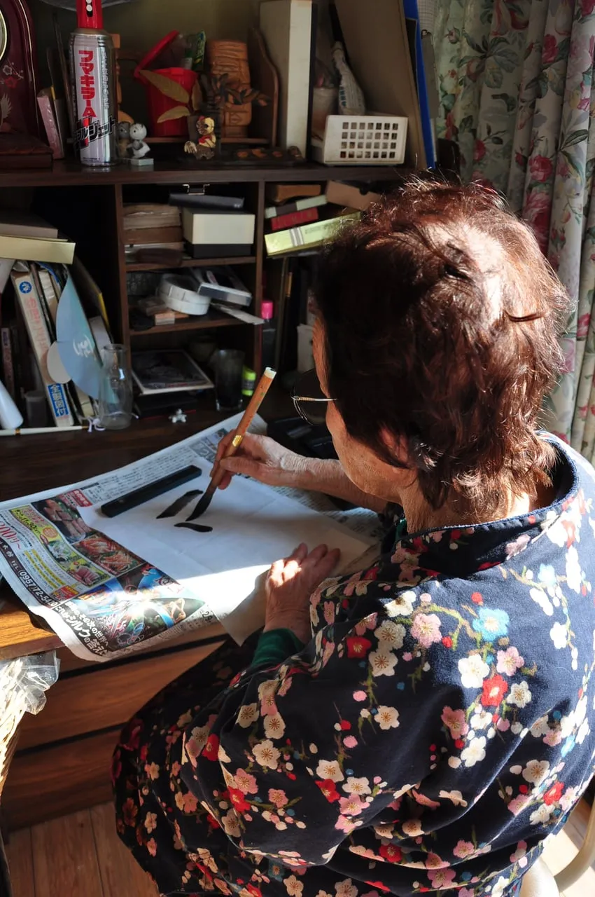Risa’s grandmother practising Japanese calligraphy with a brush at her desk.